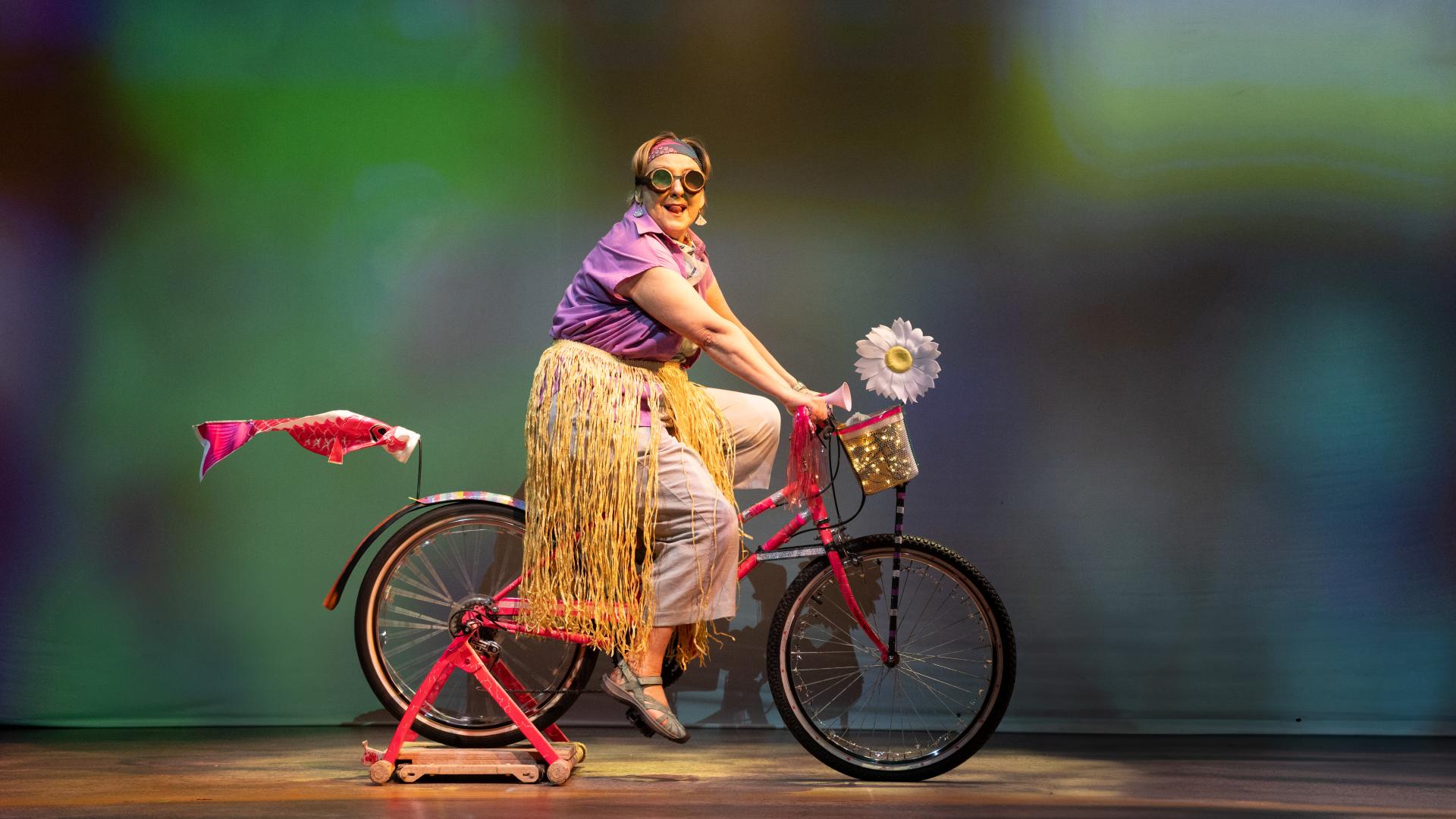 A female performer "rides" a bicycle onstage dressed in googles and a hula skirt over a purple blouse and capris.