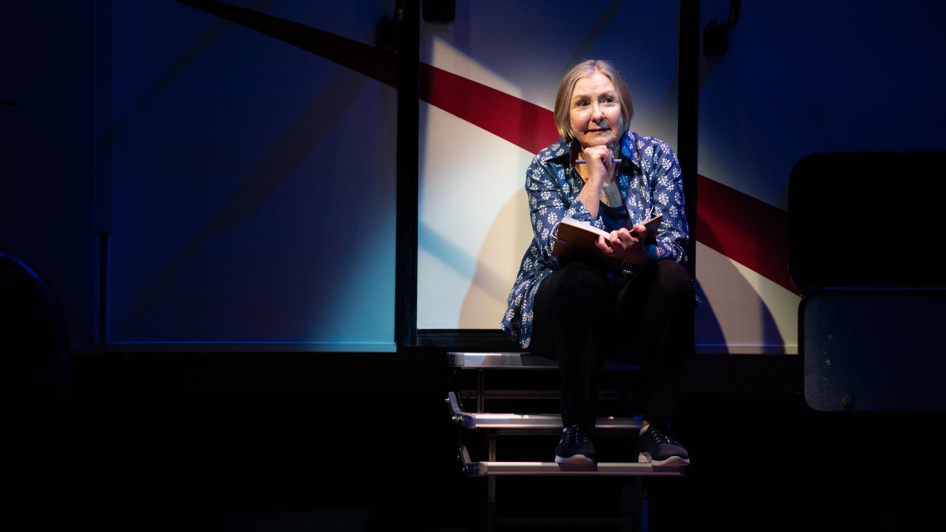 A female performer sits under a stage spotlight on the steps of a recreational vehicle while pondering a notebook she holds.