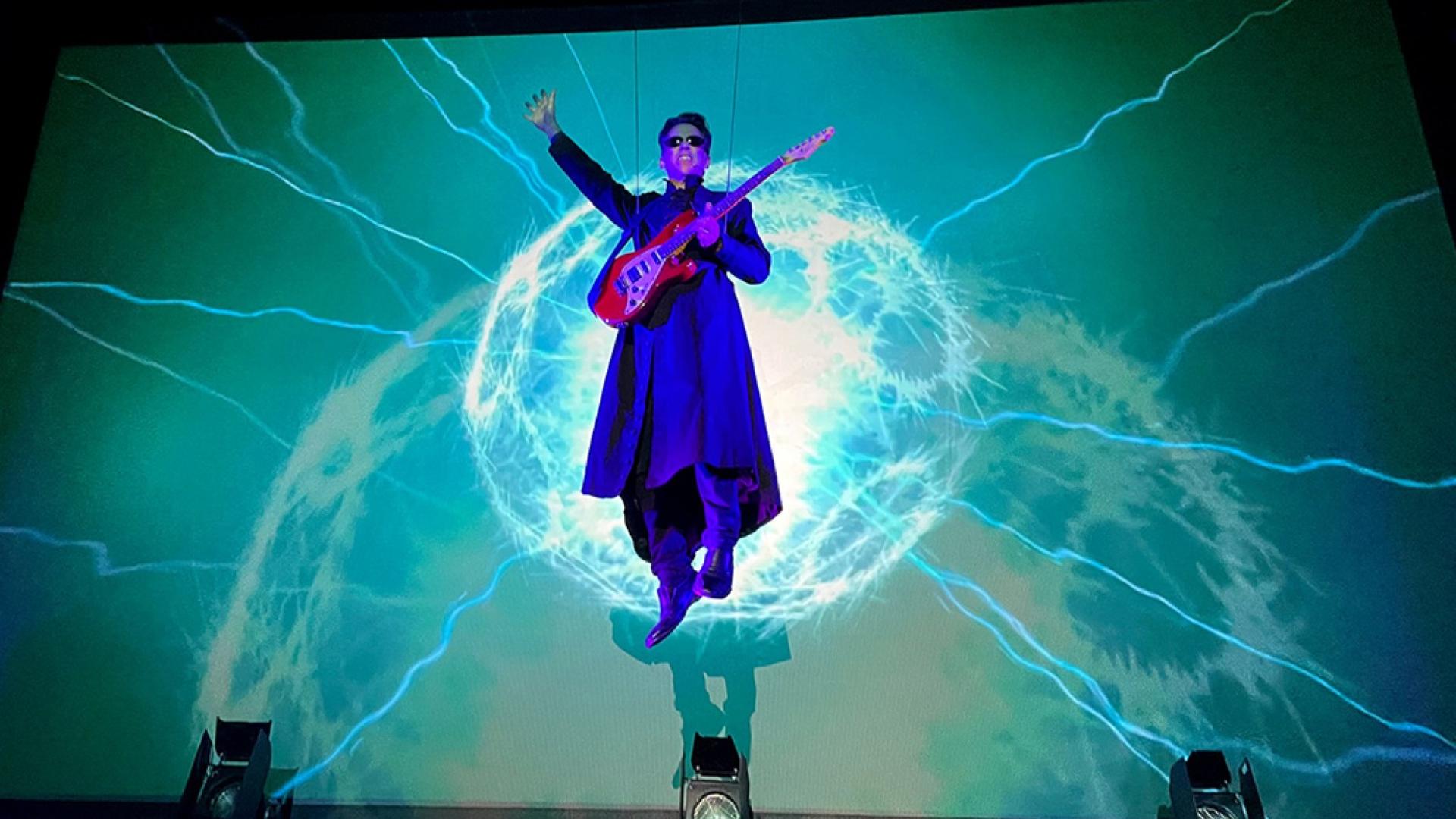 A man lit in purple rises above a stage to play a guitar while cyan lightning dances on the stage backing behind him.