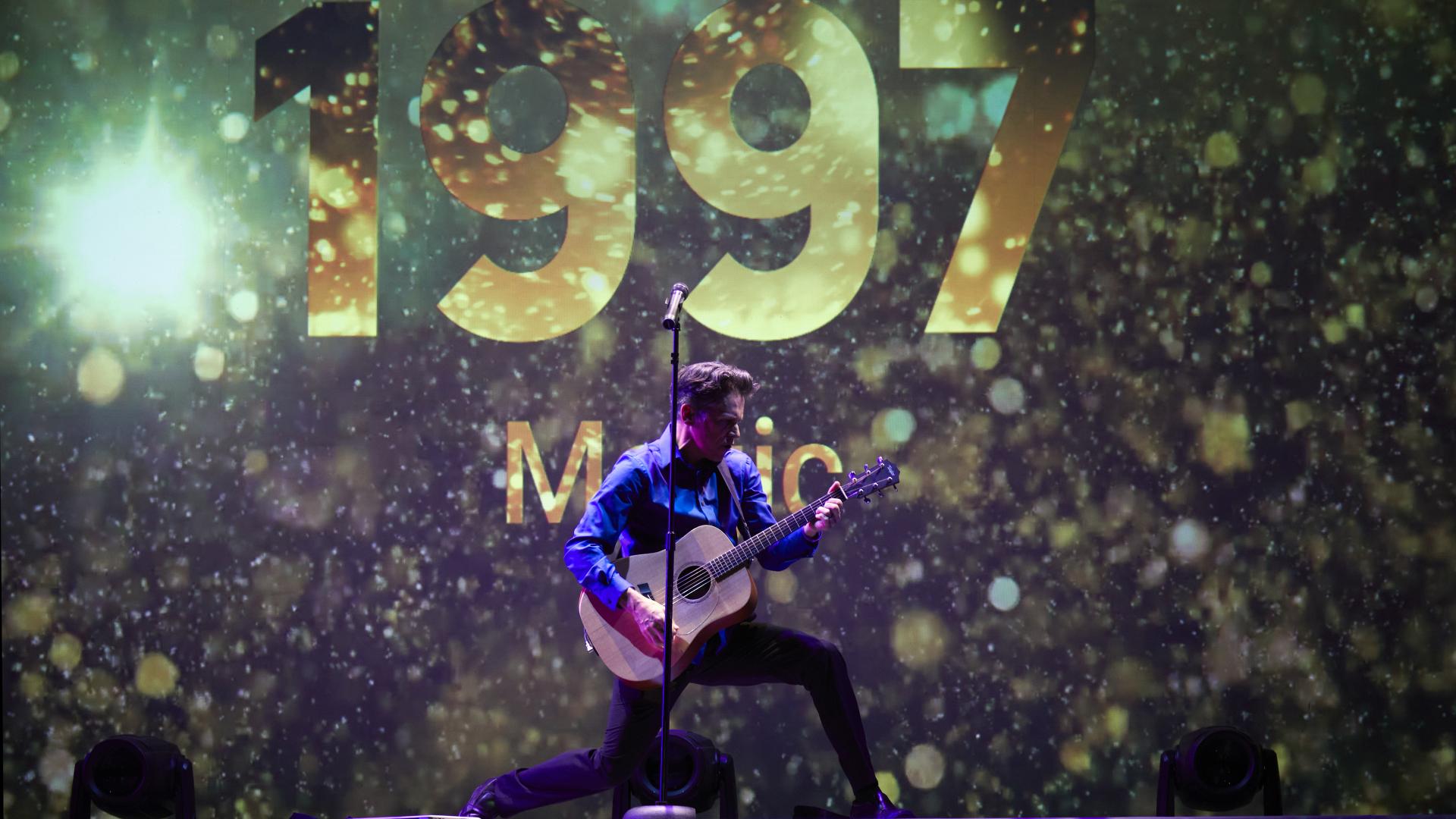A man playing a guitar kneels dramatically before a screen that proclaims 1997 in gold numerals.