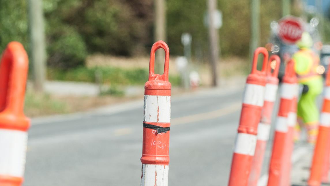 A row of orange pylons lines a roadside work area where workers in high visibility equipment are directing traffic.