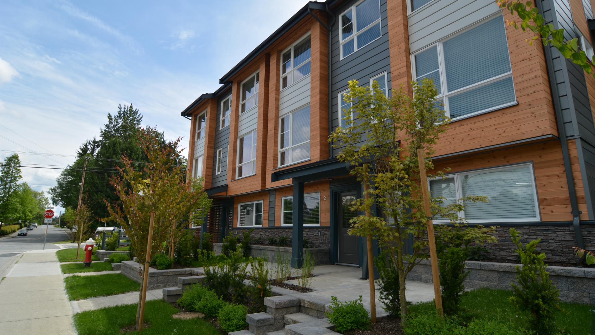 A brown and black apartment building sits behind a boulevard full of young trees next to the sidewalk.