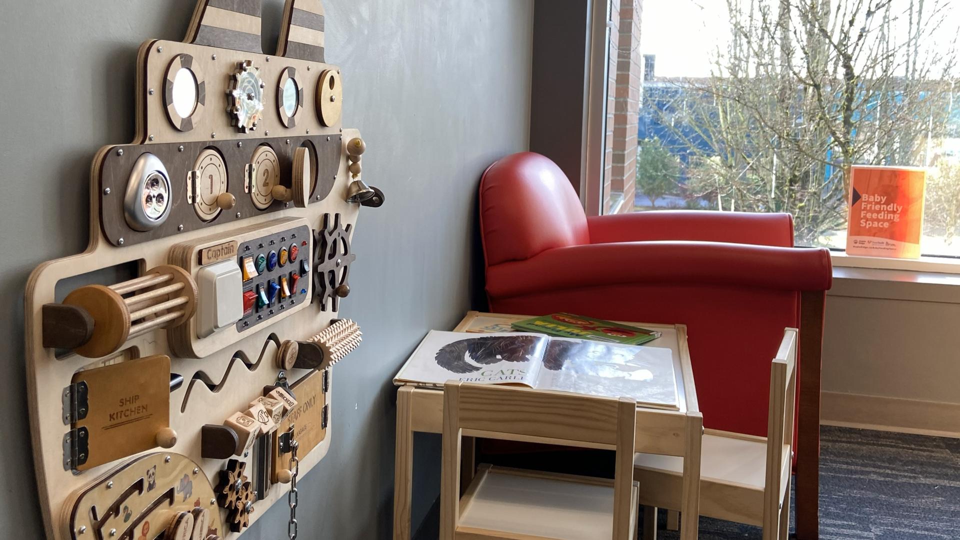 A smooth red armchair sits next to the window, with a child-sized table and chairs and wall-mounted wooden play apparatus beside it.
