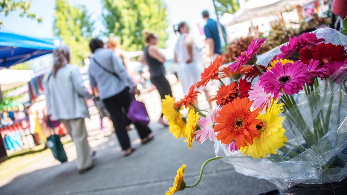 A bright bouquet of gerbera daisies, with shoppers at an outdoor market in the background.