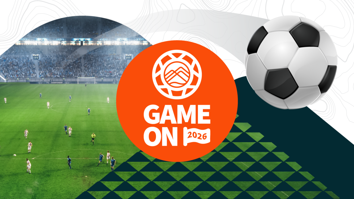 A soccer ball flies out from a field full of players, next to the Game On 2026 Logo.