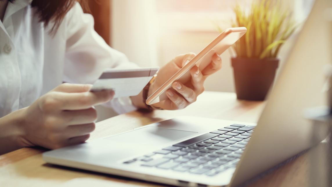 A woman holds a credit card in one hand and a phone in the other as she references a laptop screen.