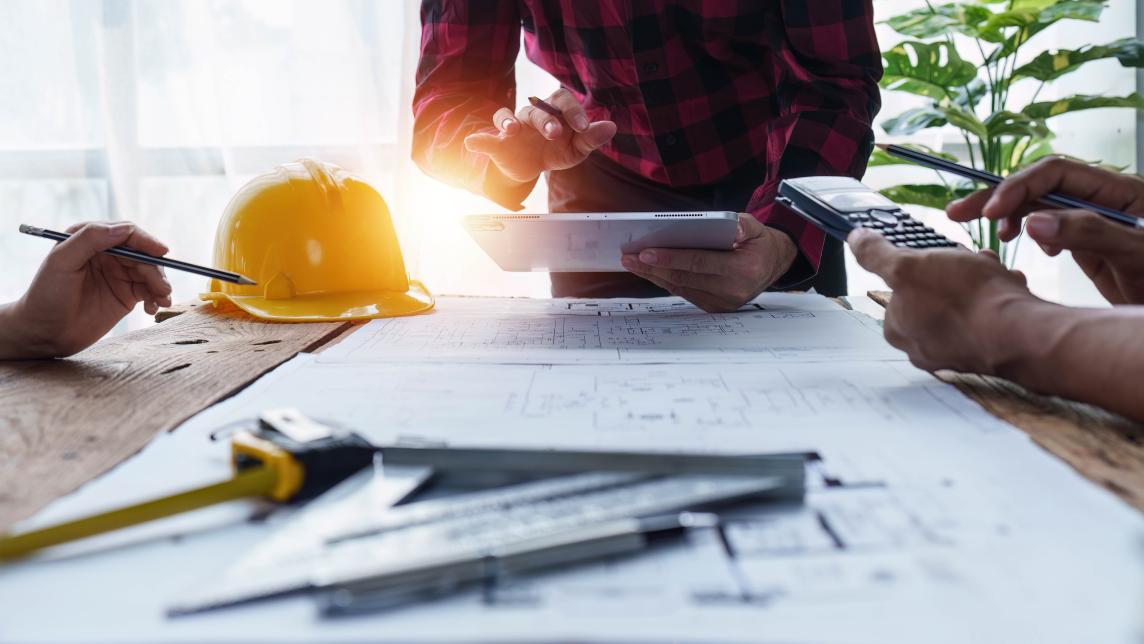 Workers lean over a table covered in construction memorabilia and housing plans. 