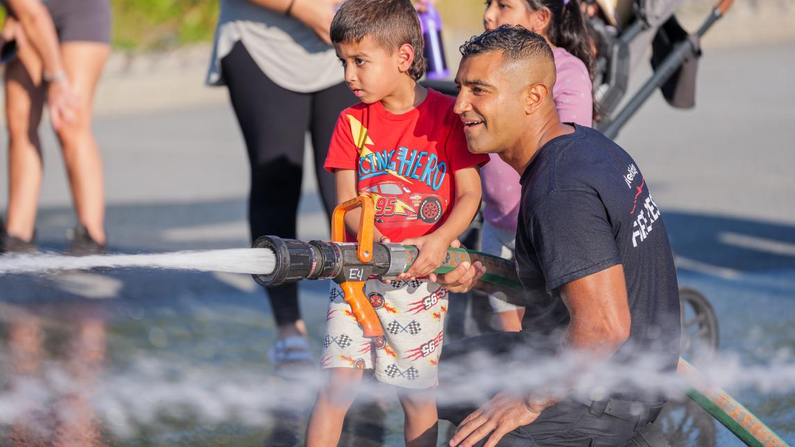 Maple Ridge firefighter helps child hold firehose at Hot Summer Nights event 