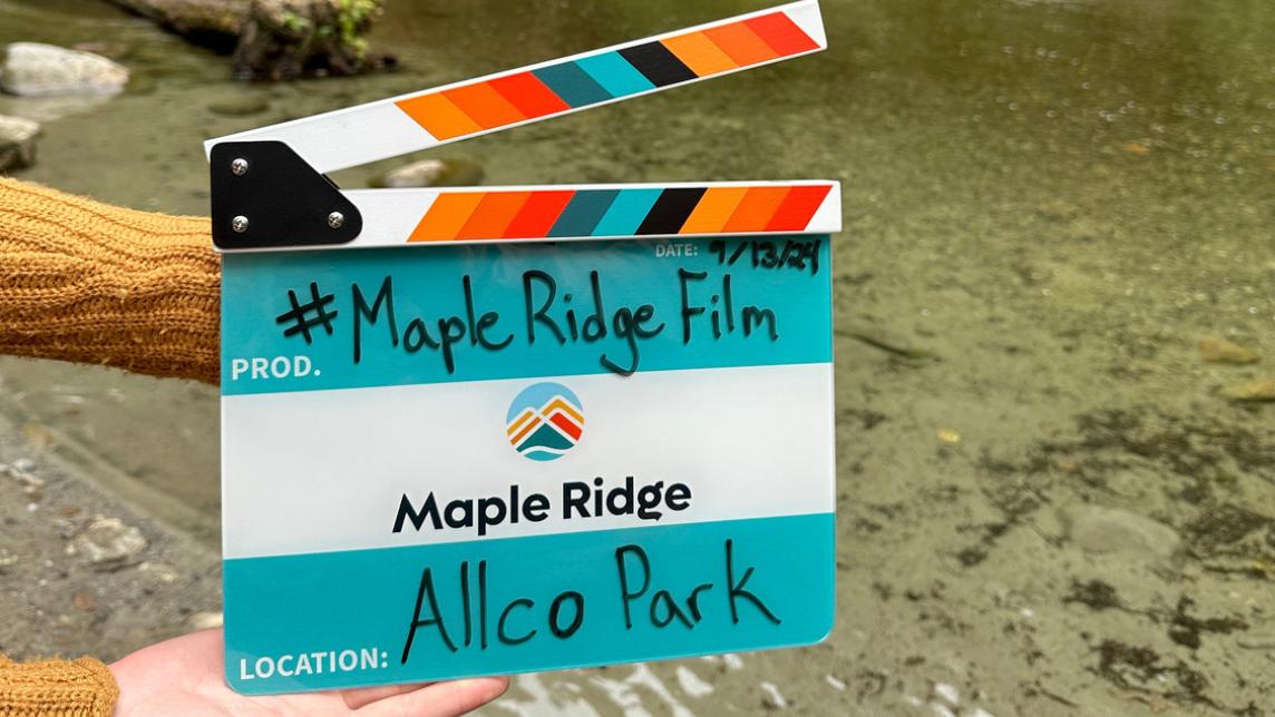 An offscreen woman holds out a City-branded film clapper with the words, #MapleRidgeFilm and Allco Park on it. The Maple Ridge logo sits in the middle.
