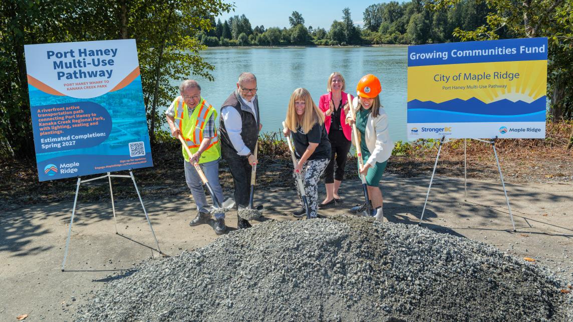 5 people (mayor, councillors, MLA) shovelling a pile of gravel beside the fraser river