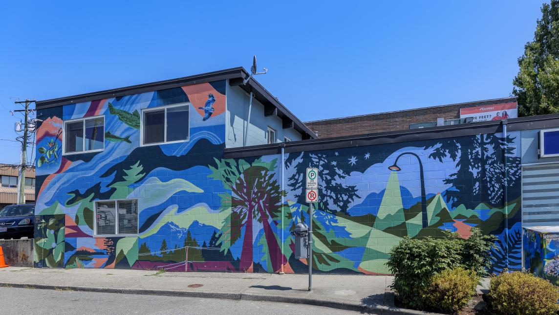 A mural with deep blues, greens and red accents covers the side of a building, depicting the Golden Ears mountains, a pine tree, and street lamp.