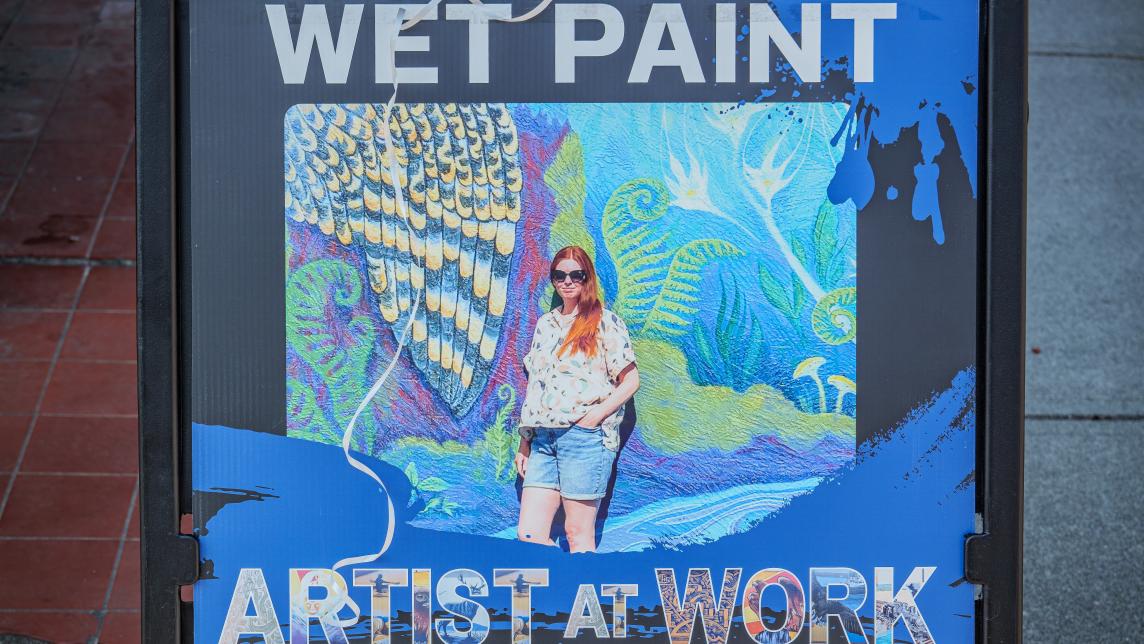 A fold-out sign warning passersby of wet paint, and the artist, Carly Bouwman, at work. It explains a bit about the Mural Collective project and its organizers and funders.