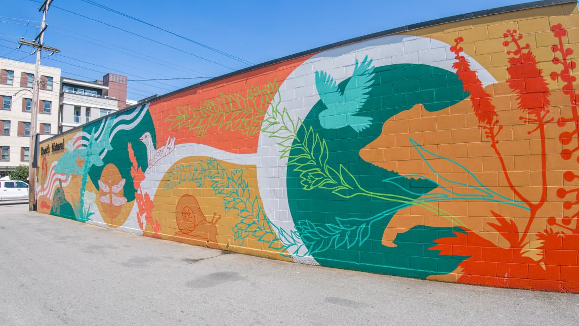 A vibrant mural in reds, teal and oranges depicts native species, such as a heron, dragonfly, snail, and owl alongside plant imagry.