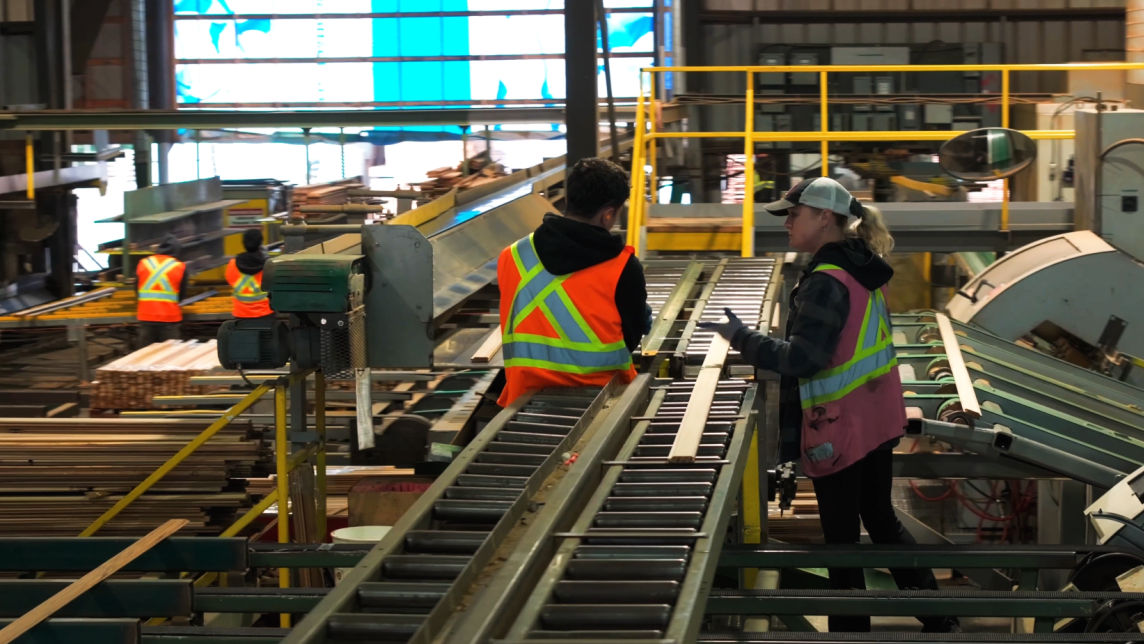 A pair of workers in high visibility vests talk next to a set of conveyor belts in a wearhouse.