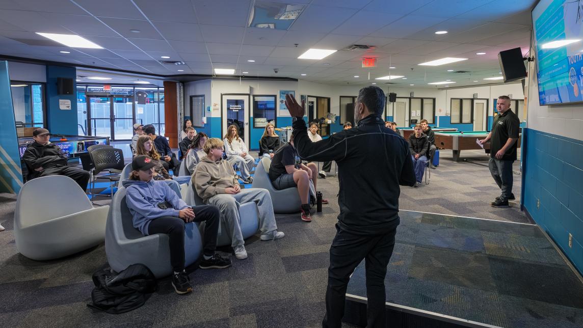 A man stands, speaking, in front of a group of grade nines seated in the Greg Moore Youth Centre on Take Your Grade Nine to Work day.