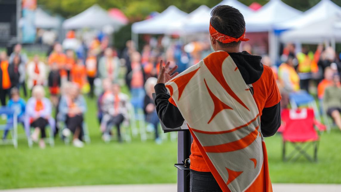 A man wearing an orange handkerchief headband, shirt, and orange-and-white sash with an indigenous pattern speaks to a crowd.