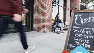 A person walks by a chalkboard "Open" sign for a store, while another person rests on a ledge checking their phone above a blue portion with text that reads "News Release Free Buy Local Workshop for Maple Ridge Businesses on April 8".