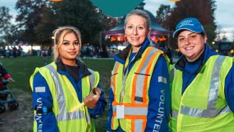 Three female volunteers in high-visibility vest smile at the camera at the Celebrate the Night Event in Memorial Peace Park. Above them is white text on a blue background that reads: "Thank You! We Are Driven By Community #NationalVolunteerWeek Maple Ridge"