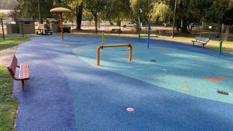 A red bench sits next to a grassy area that border the Kinsman Spray Park at Maple Ridge Park. The blue splash pad is covered with yellow and green water apparatuses.