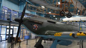 A plane with a nose propeller sits under a ceiling-mounted biplane in a museum exhibit.