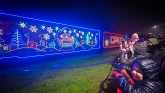 A line of onlookers bundled in winter gear watch as the CPKC Holiday Train passes by, lit up in a neon display.