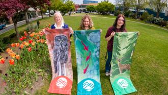 The three artists behind the winning 2026 Banner designs stand in Memorial Peace Park with the completed banners.