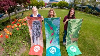 The three artists behind the winning 2026 Banner designs stand in Memorial Peace Park with the completed banners.