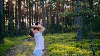 A young boy in a forest cranes his neck to look straight up at the treetops with binoculars.