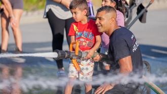 Maple Ridge firefighter helps child hold firehose at Hot Summer Nights event 
