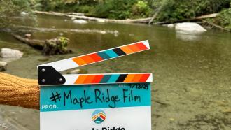 An offscreen woman holds out a City-branded film clapper with the words, #MapleRidgeFilm and Allco Park on it. The Maple Ridge logo sits in the middle.