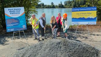 5 people (mayor, councillors, MLA) shovelling a pile of gravel beside the fraser river