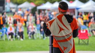 A man wearing an orange handkerchief headband, shirt, and orange-and-white sash with an indigenous pattern speaks to a crowd. 