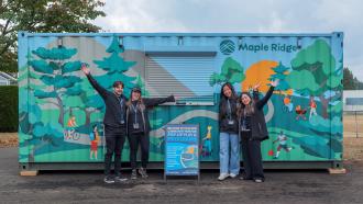 Four City of Maple Ridge Staff stand in front of a "Rec Box," a modified shipping container painted with an image of a sunny park.