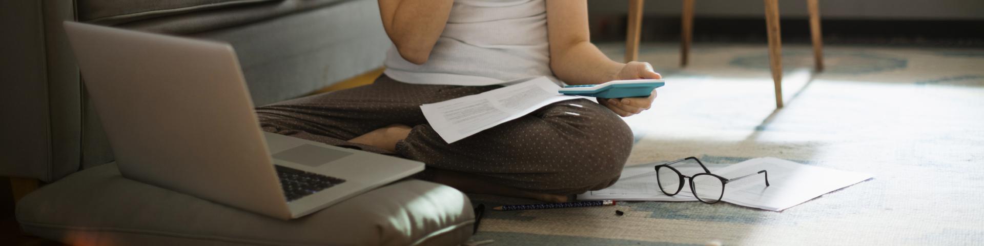 A woman sits cross-legged on the floor, her glasses beside her as she evaluates paperwork next to a computer.