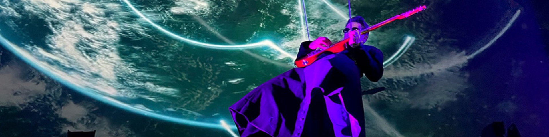 A man lit in purple leans back to play a guitar while cyan lights dance on the stage backing behind him.