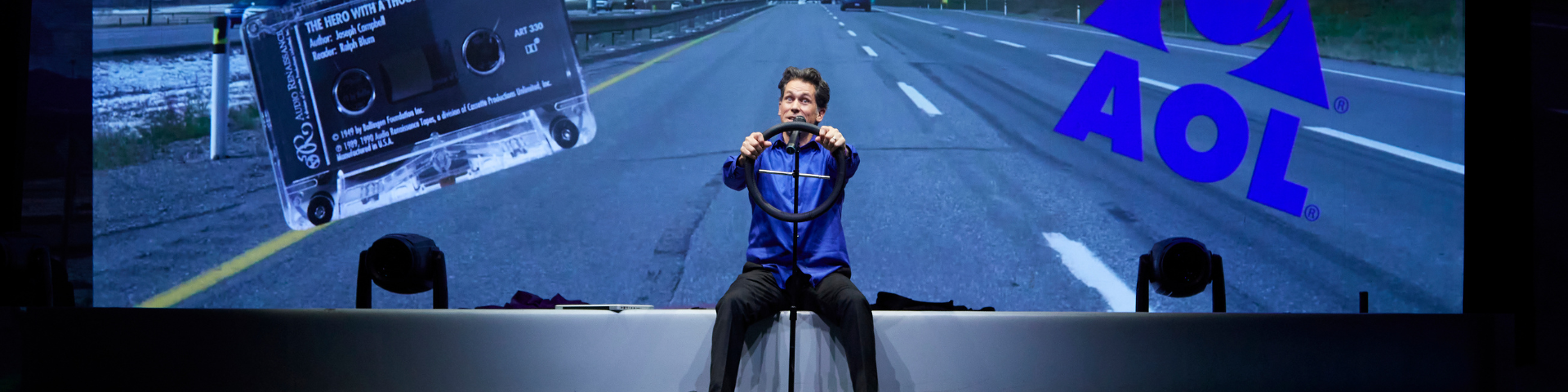 A man mimes driving a car onstage while a projector shows the road slipping away behind him.