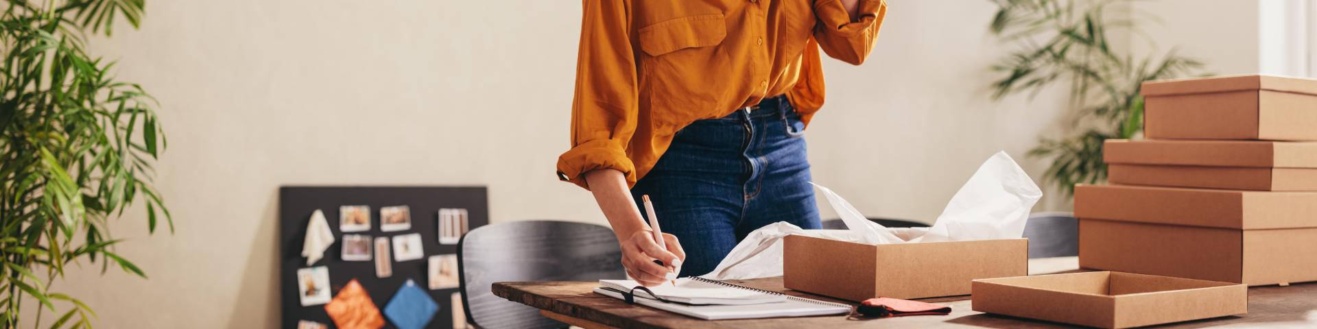 A woman reaches down to scribble notes on a piece of paper while talking on the phone.
