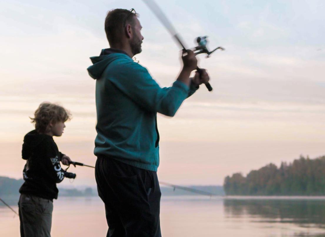 A young boy and his father cast their fishing rods into a lake in the early morning light.