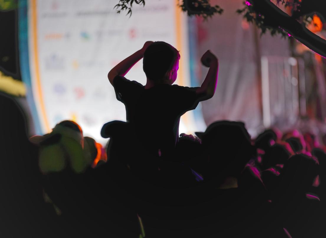 A child holds up his hands as he sits on the shoulders of their guardian, watching the stage at the 2024 Out Neck of the Woods event in Memorial Peace Park.
