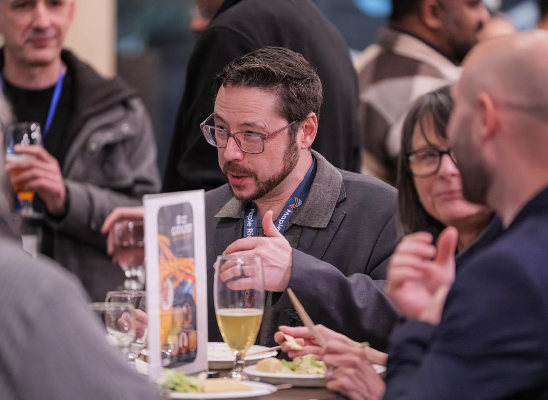 A man gesticulates as he talks to a small crowd of people around a table.
