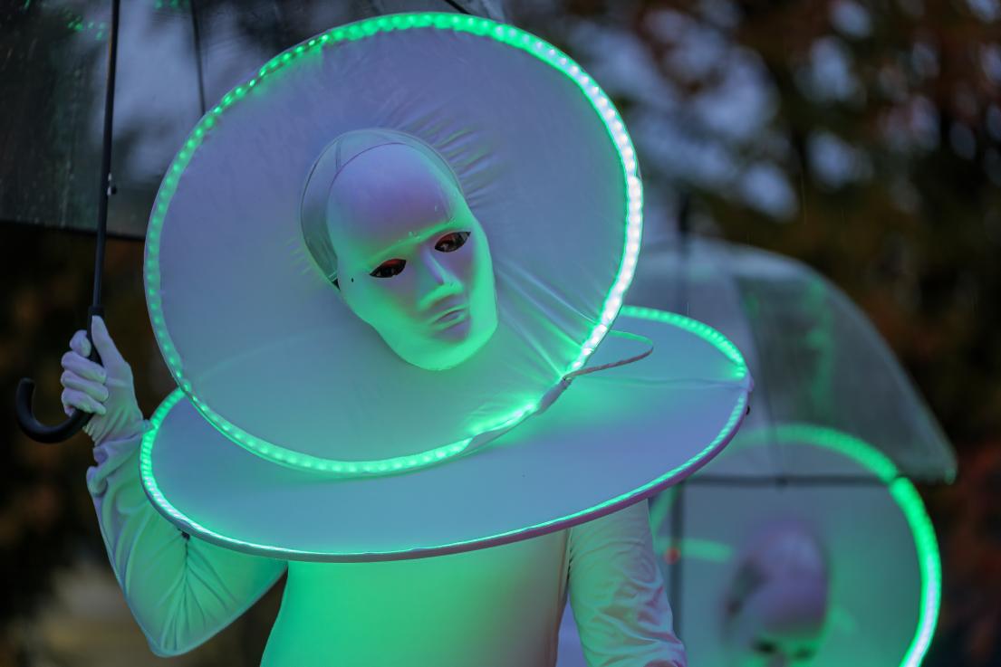 A performer wears a white costume with inset lights to make them appear alien.
