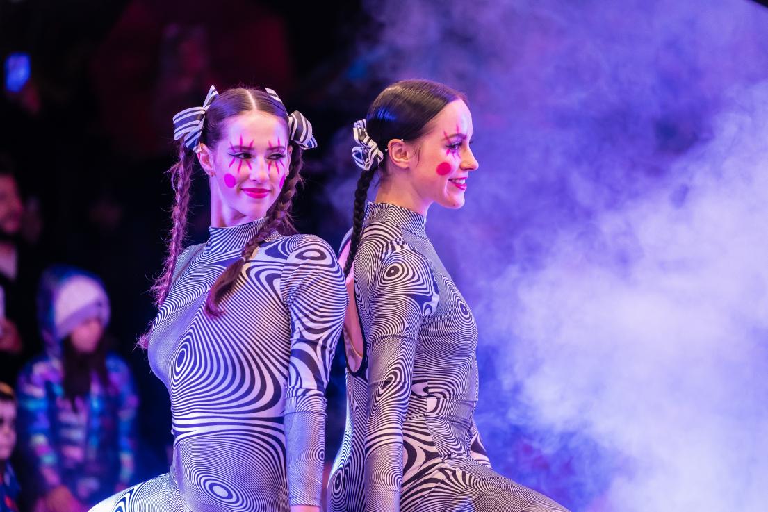 Two female performers sit back to back against artificial smoke, wearing striking white and black striped one-piece suits.