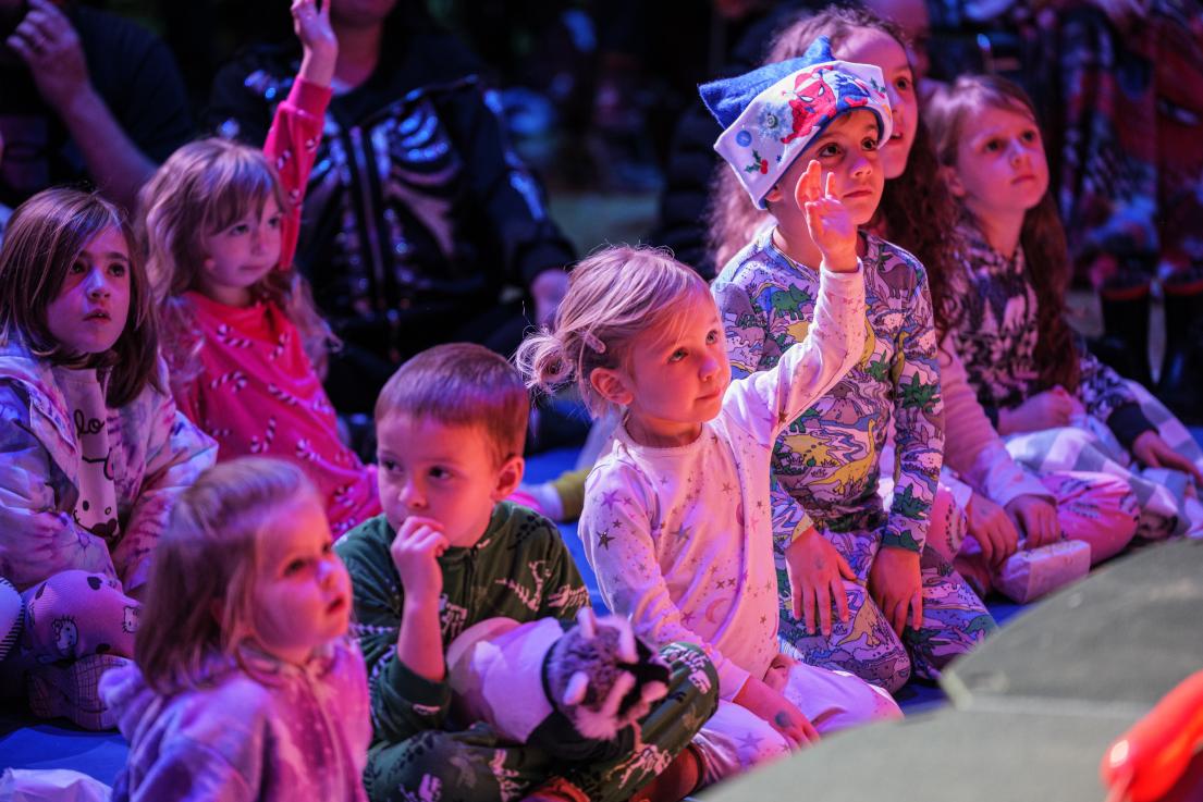 A crowd of kids wearing pyjamas raise their hands to volunteer as they watch a performer.