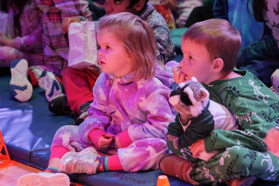 Two young children in pyjamas sit on mats as they watch a performer.