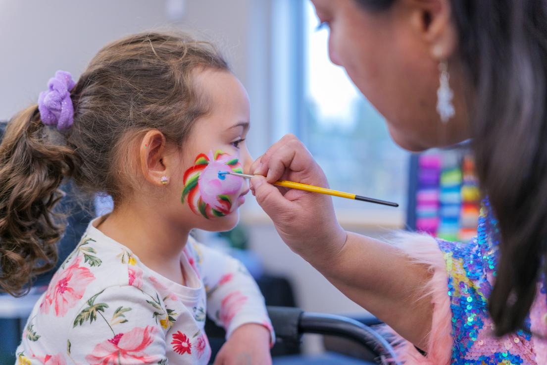 A child turns her face to the side so an artist can finish painting a prink unicorn with rainbow mane on her cheek.