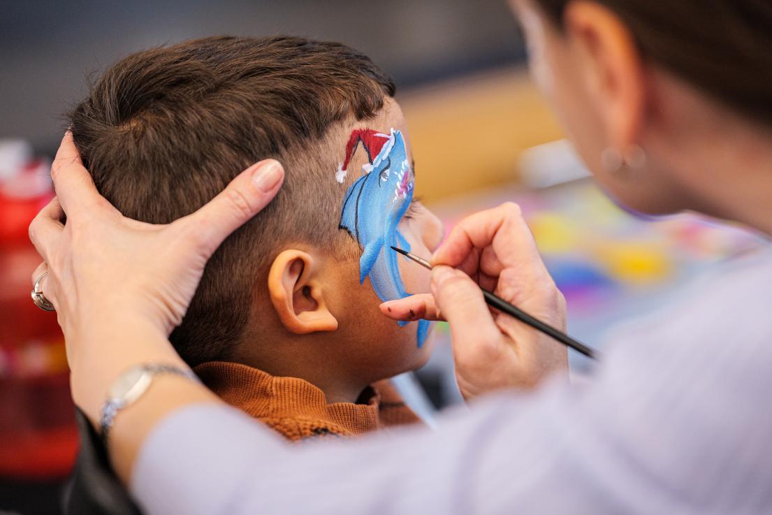 An artist carefully paints a shark wearing a Santa-hat on a young boy's face.