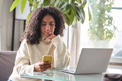 woman looking at her mobile phone sitting at a desk with a laptop on it