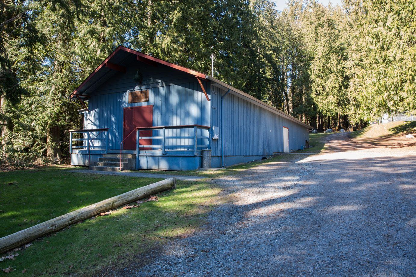 A narrow and long blue, wood sided building. It sits in a wooded area next to a gravel parking lot.