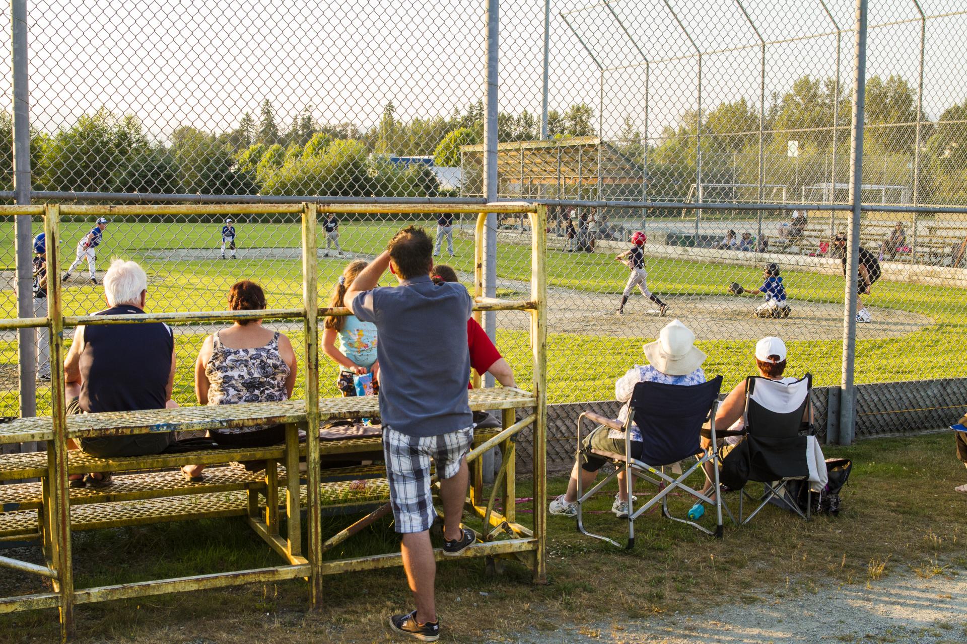 A crowd of people watch a baseball game from the other side of the fence on bleachers and lawn chairs at the Albion Sports Complex.
