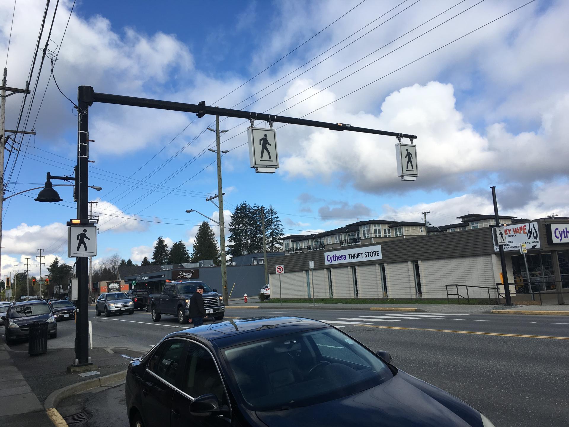 A car is parked next to a crosswalk on Dewdney Trunk Road. A pedestrian crosses the street while the cars in the road wait.